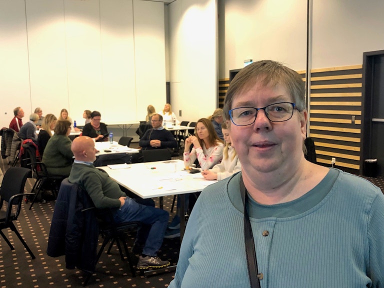 Bilde av Vigdis Løbach inne på konferansesal med mennesker på diverse bord i bakgrunnen