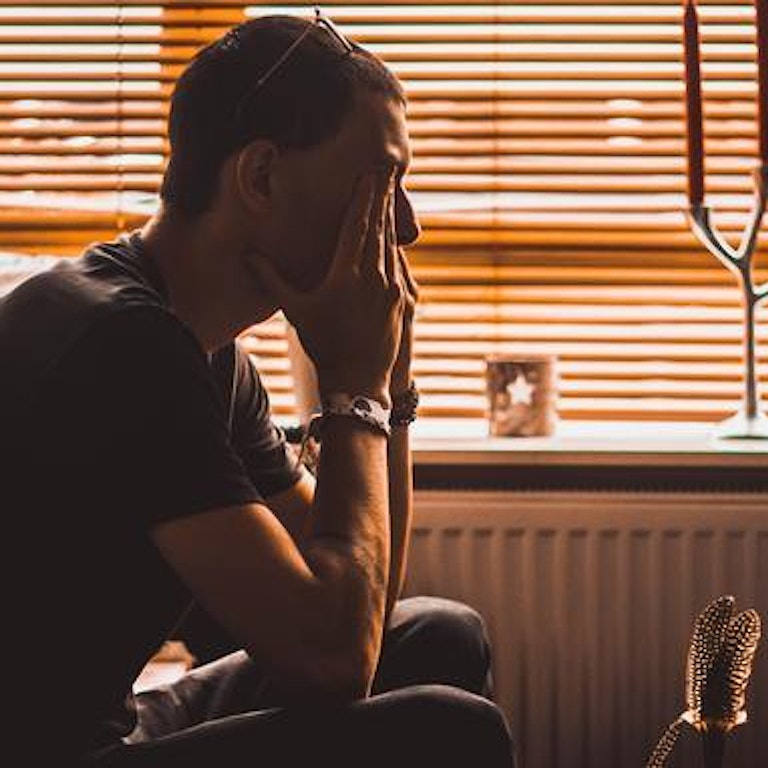 En mann sitter innendørs med gardinene trukket for. Han hviler ansiktet i hendene. (Foto: Christopher Lemercier via unsplash)