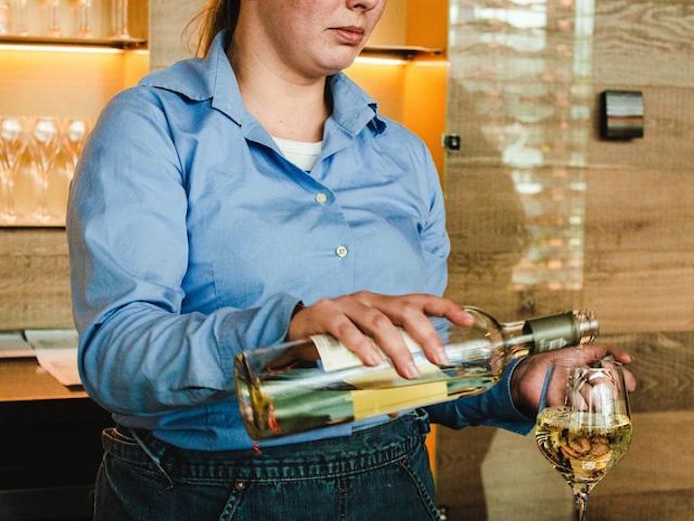 En kvinne står bak en bardisk og heller vin i et glass. (Foto: Louis Hansel via unsplash)