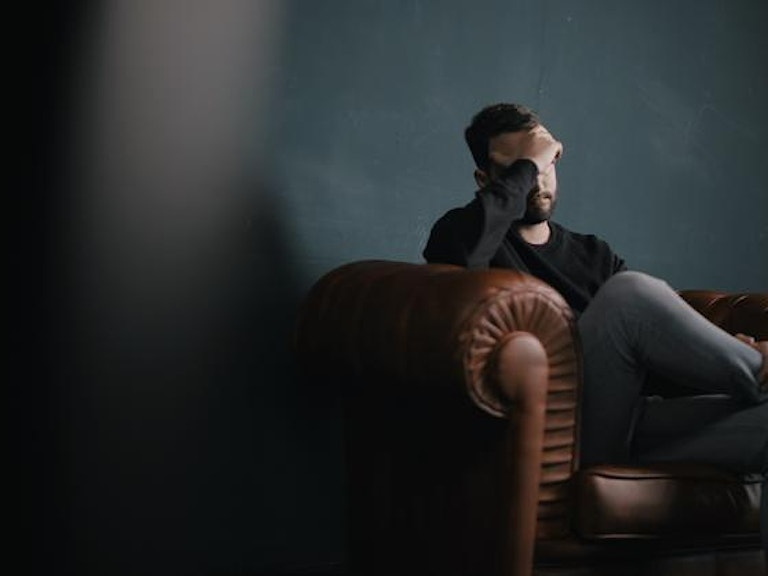 En mann sitter i en sofa med hodet lent i hånda. Øynene hans er skjult.(Foto: Nik Shuliahin via unsplash)