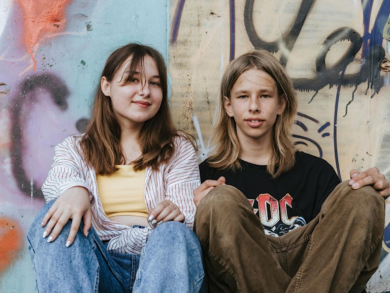 Foto av to ungdommer sitter foran en vegg med grafitti. De ser inn i kameraet og smiler.