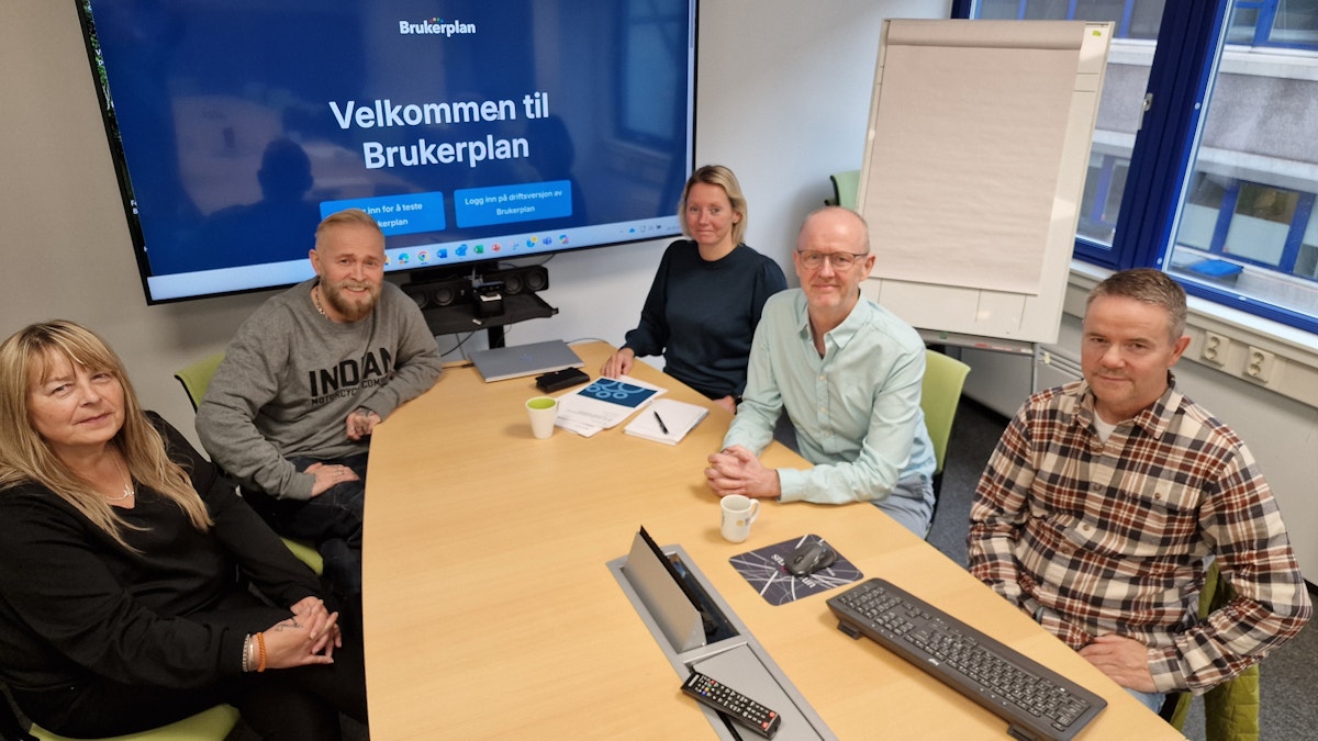 En gruppe fra KORUS og kommunen sitter rundt et stort langbord foran en skjerm som viser tittelen "Velkommen til Brukerplan".
