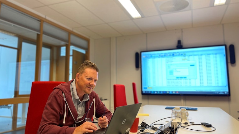 Sven Gustafsson fra KORUS Stavanger sitter ved et langbord på et kontor med en laptop. På storskjerm foran i rommet ser vi flere tall.