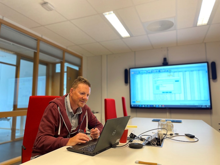 Sven Gustafsson fra KORUS Stavanger sitter ved et langbord på et kontor med en laptop. På storskjerm foran i rommet ser vi flere tall.