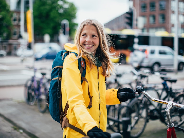 En ung kvinne står og holder på en sykkel. Hun har på seg en knall gul jakke. Hun ser litt til venstre for kameraet og smiler bredt.