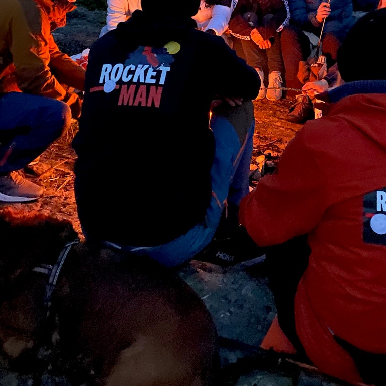 Mennsker som sitter rundt et bål. En har en genser med påskriften "Rocket Man"