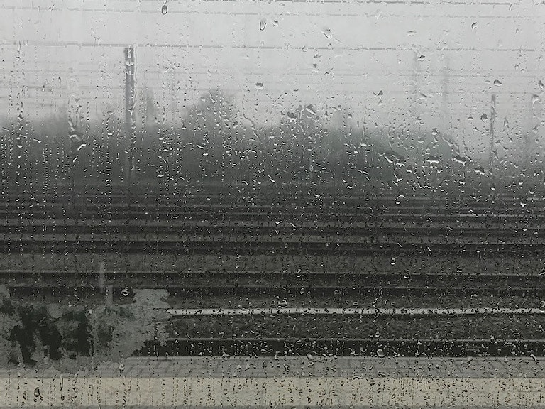 Illustrasjonsfoto. Viser et togvindu vått av regn. Fokus på vindusflaten. Uskarp bakgrunn cirka øverste tredjedel himmel, resten et stasjonsområde.