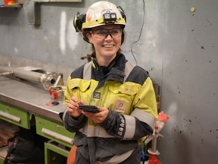 Sara fra Bilfinger er kledd i full arbeidsuniform. Gul arbeidsdress, hjelm, hoerselvern og bryller. Hun taster paa mobilen.