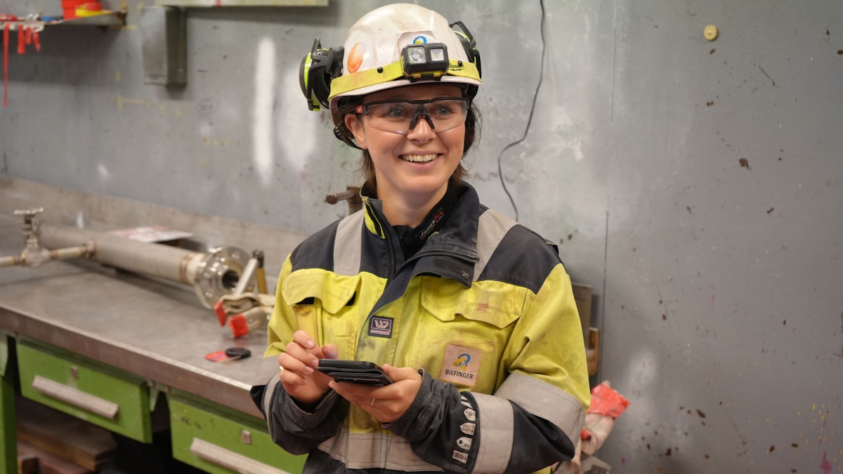 Sara fra Bilfinger er kledd i full arbeidsuniform. Gul arbeidsdress, hjelm, hoerselvern og bryller. Hun taster paa mobilen.