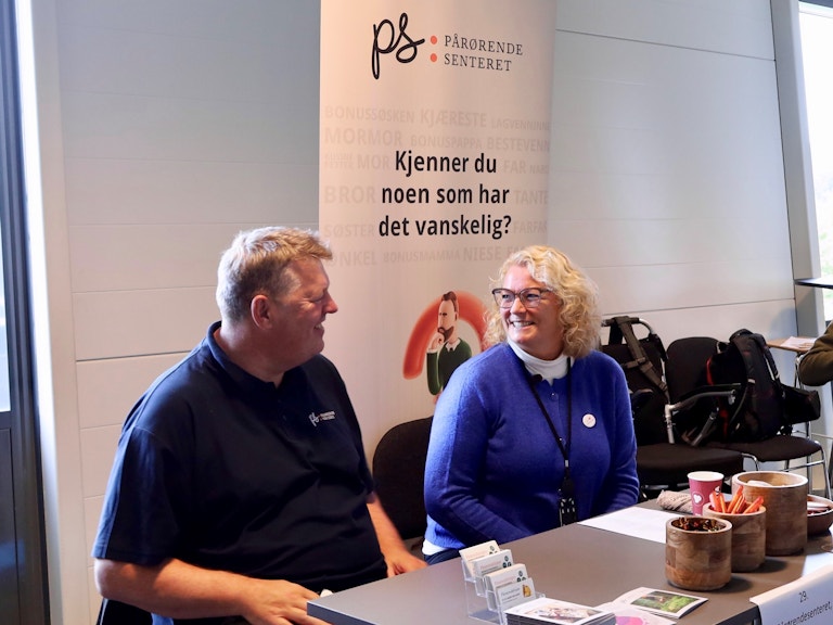 Bildet viser to personer som sitter bak et bord. De smiler til hverandre, og på en stor plakat bak dem står det «Pårørendesenteret. Kjenner du noen som har det vanskelig?»