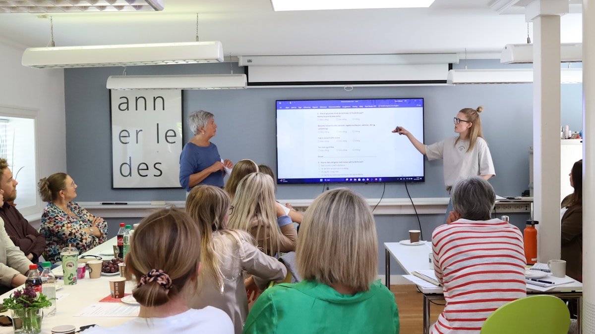 Bildet viser to kvinner som holder en presentasjon. De står på hver sin side av en stor skjerm, og kvinnen til høyre peker på skjermen. Foran dem ser vi bakhodet på rundt ti mennesker som følger med på det de sier.