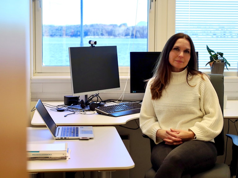 Portrettfoto av Marte Kristin Helle. På bildet ser vi henne sittende ved siden av en pult. Bak henne er det to skjermer, og ved siden av henne er det en laptop. Hun sitter vendt mot kameraet- med hendene i fanget og et mykt smil.