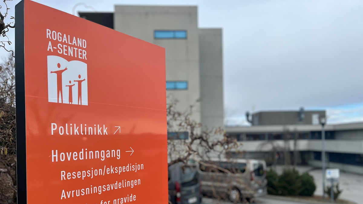 Bilde av et stort, oransje skilt som står på parkeringsplassen til Rogaland A-senter. Skiltet er i fokus, mens bakgrunnen er uskarp. På skiltet ser vi A-senterets logo, og nedover på skiltet pekes det vei til poliklinikken, hovedinngangen, resepsjonen, avrusningsavdelingen, avdeling for gravide og småbarnsfamilier og behandlingsavdelingen.