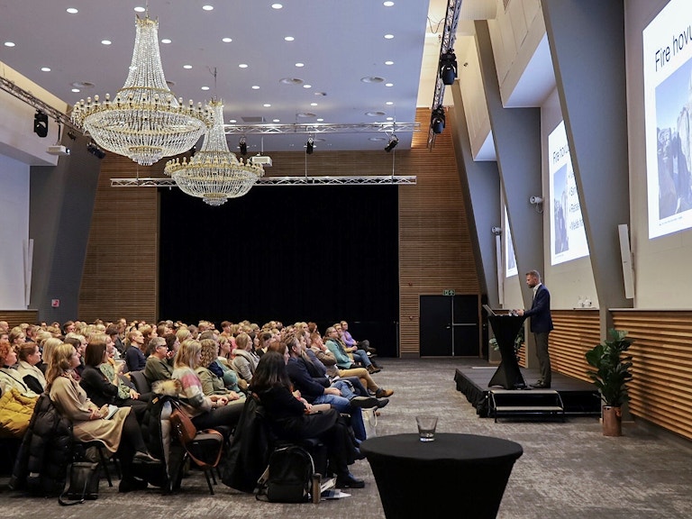 Bildet viser en stor konferansesal med to store lysekroner i taket. Bildet er tatt fra siden. Til høyre ser vi Bent Høie, Statsforvalteren i Rogaland, som står på et podie og snakker til forsamlingen. Bak ham er tre storskjermer, og rett overfor ham sitter publikum.