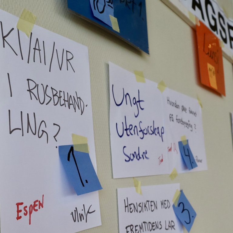 Nærbilde av ark som henger på en vegg. På arket nærmest oss står det "KI/AI/VR i rusbehandling?" og på neste ark står det "Ungt utenforskap". Det henger flere ark bortover veggen, men disse er ikke i fokus og derfor ikke mulige å lese. På alle lappene er det festet fargerike post it-lapper med et tall på.