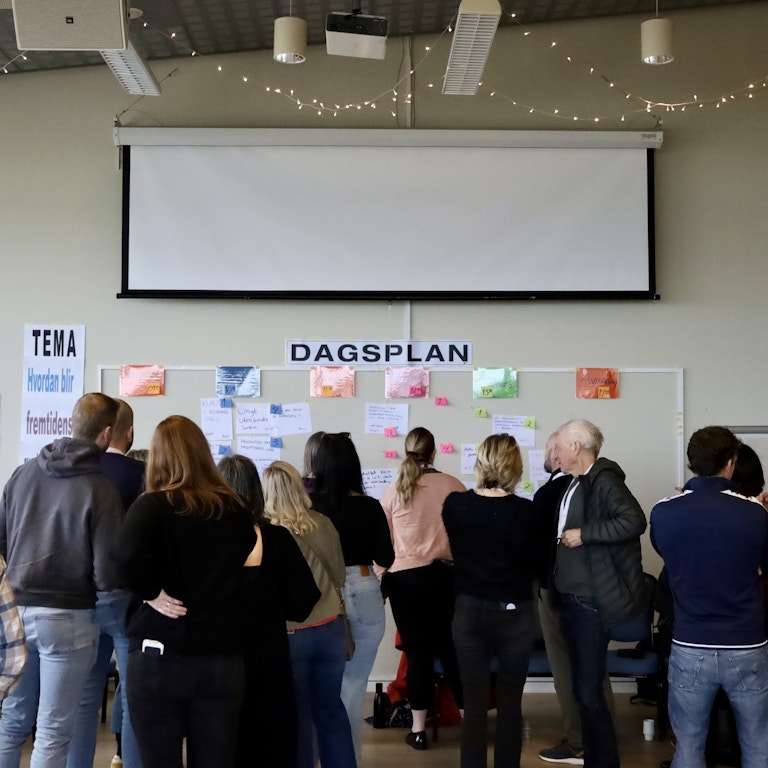 Bildet viser et stort rom fylt av mennesker. Alle menneskene står vendt mot en tavle, hvor det henger en stor lapp hvor det står "Dagsplan"