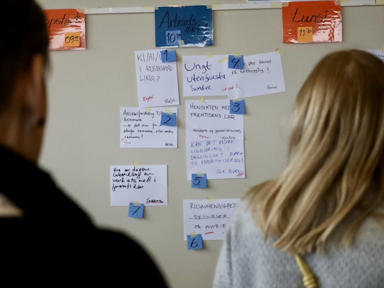 Vi ser silhuetten av to personer bakfra. Mellom dem ser vi flere ark som henger på en tavle, med blå post it-lapper festet på hver av dem. Arkene er for langt unna til at vi klarer å lese hva som står, men det ser ut som om de to personene prøver å lese hva som står.