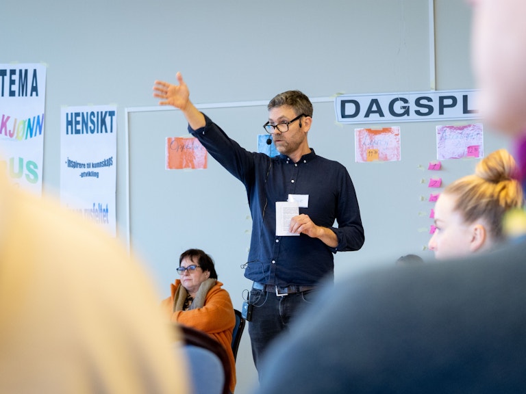 Bilde av Tore Berge. Han står i midten av en forsamling. Det ser ut som om han er midt i en setning, og han bruker den høyre armen til å peke på noe eller vise vei.