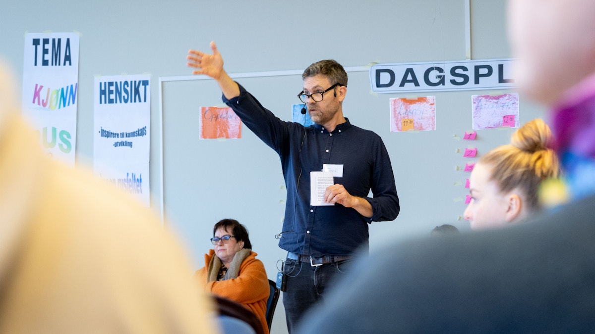 Bilde av Tore Berge. Han står i midten av en forsamling. Det ser ut som om han er midt i en setning, og han bruker den høyre armen til å peke på noe eller vise vei.