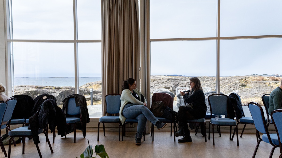 Bildet viser to kvinner som sitter sidelengs og snakker med hverandre. Rundt dem er det tomme stoler, og bak dem er det to store vinduer med utsikt over Solastranda.