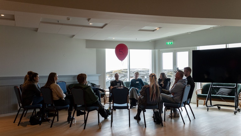 Ti-tolv personer sitter på stoler i en sirkel, og en stor, rød heliumballong henger i en av stolene. Bildet er tatt i motlys, så det er vanskelig å se personene. Bak dem ser vi svaberg, sjø og himmel gjennom vinduet.