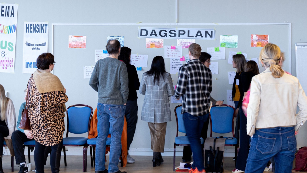 Bildet viser ryggen på cirka ti personer. Alle ser opp mot en stor tavle hvor det står skrevet «Dagsplan», og under det er det hengt opp ulike diskusjonsforslag på A4-ark. Det er ikke mulig å lese hva som står på dem fra denne avstanden, men vi ser at de henger der. Ved siden av tavlen henger det en plakat hvor det står «Tema: Kjønn, rus og avhengighet».