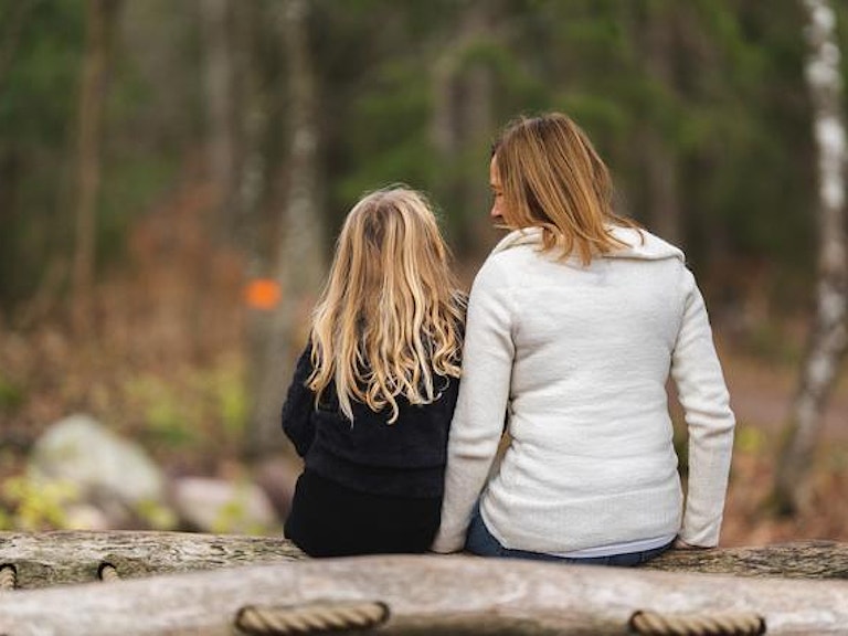 En kvinne og en ung jente sitter med ryggen til på en tømmerstokk i skogen og prater sammen. Foto: Jonas Ingstad