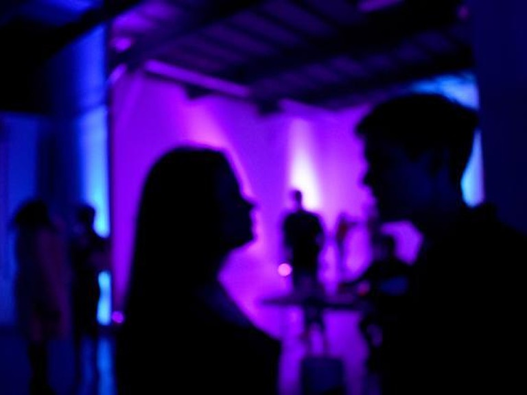 To personer som ser på hverandre inne i et mørkt lokale med lilla lys. i bakgrunnen er det flere personer