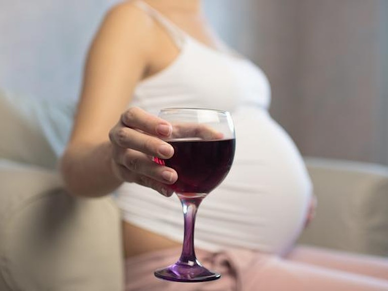 En gravid kvinne sitter i en stol med et glass rødvin i hånden. Vi ser ikke ansiktet hennes. (Foto: Istock)