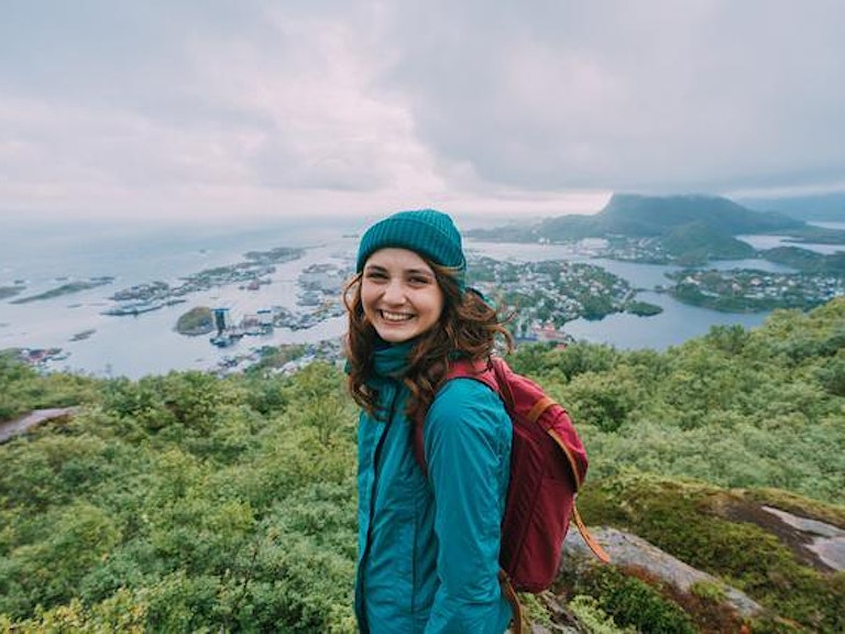 En ung jente står på toppen av et fjell. Det er flott utsikt over havet. Hun er inn i kamera og smiler fornøyd.