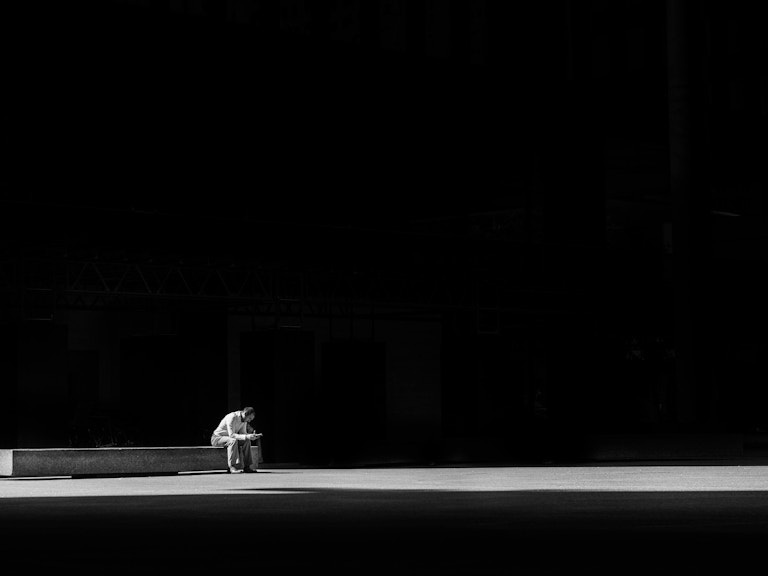 Mann sitter alene på en benk i et enormt mørkt rom. (Foto: Matthew Henry, Unsplash)